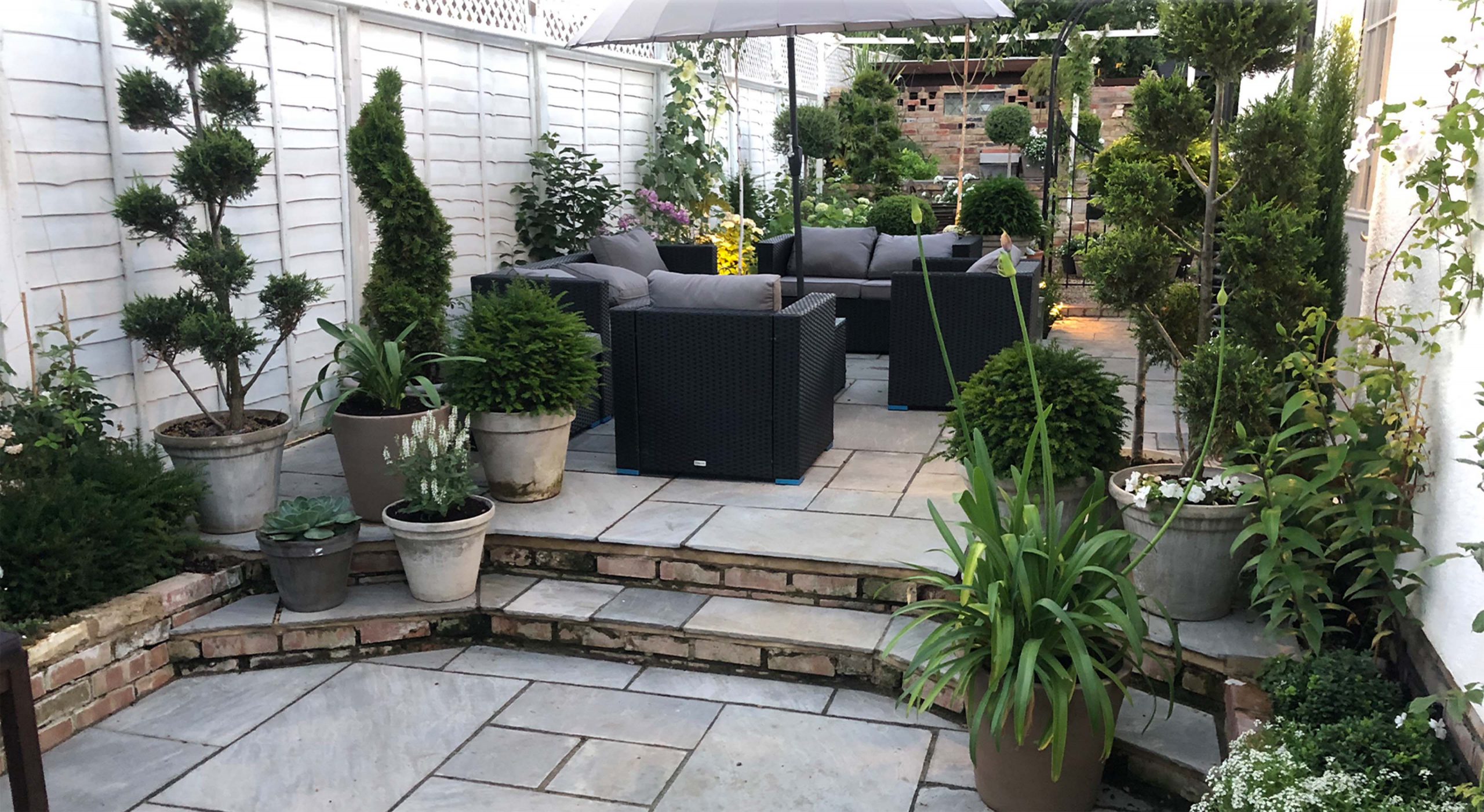 A multi-tiered tiled garden patio filled with a variety of potted plants and a seven-seater rattan furniture set designed by Willow Alexander