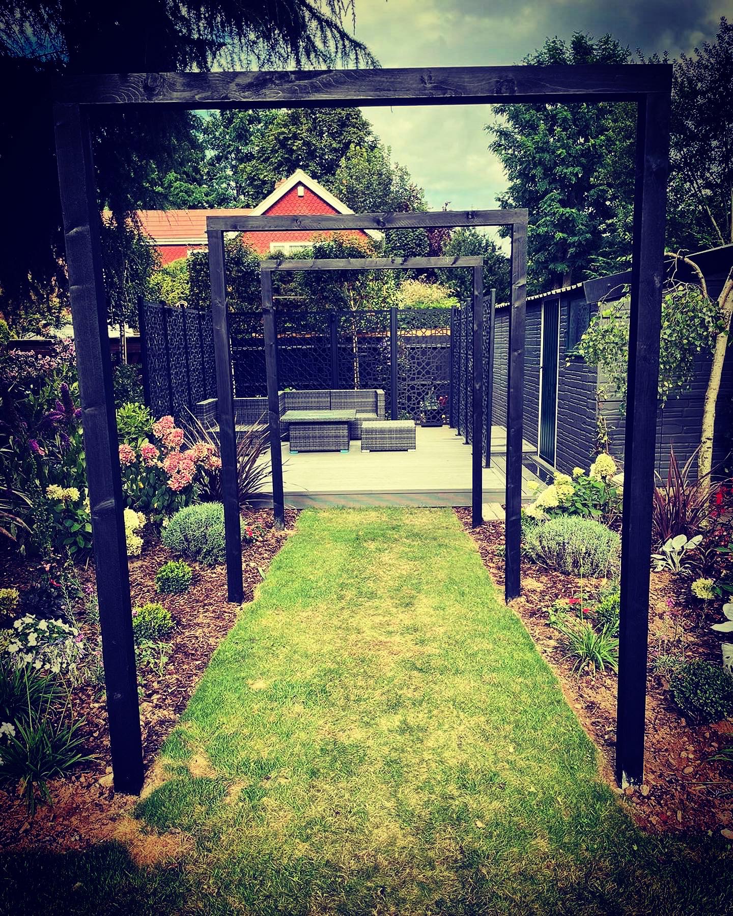 Black metal garden arches with a well-maintained garden pathway leading to a stone patio with rattan garden furniture
