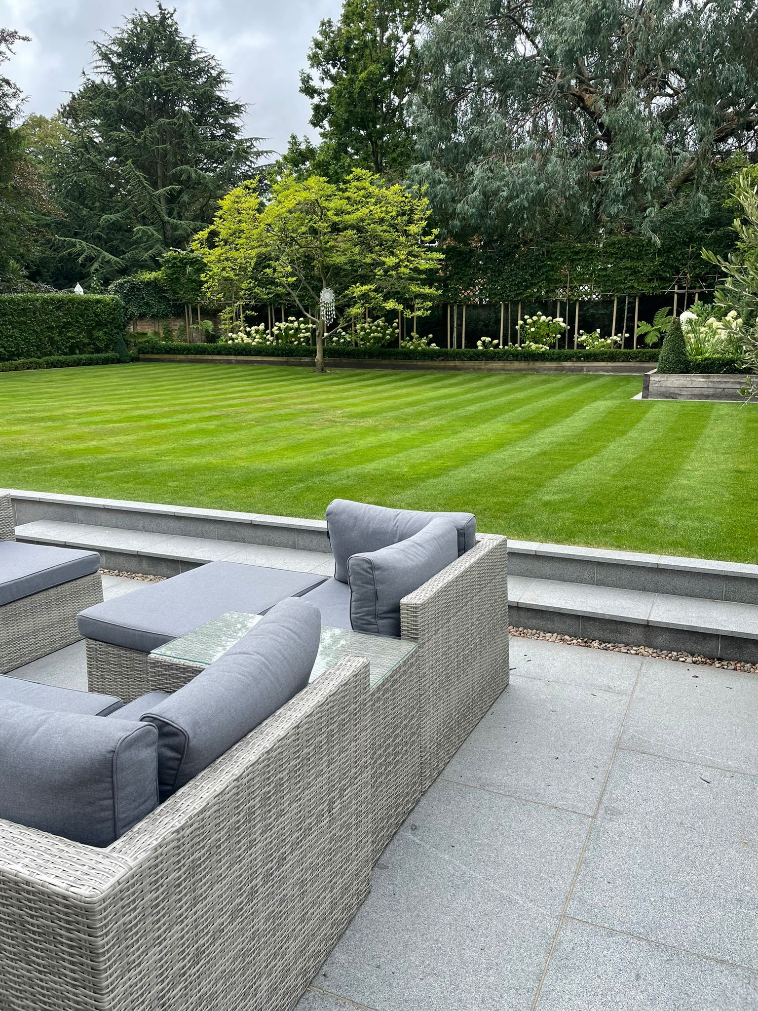 A tiled stone patio with a light-grey rattan garden furniture set with steps leading to a well-mowed garden lawn