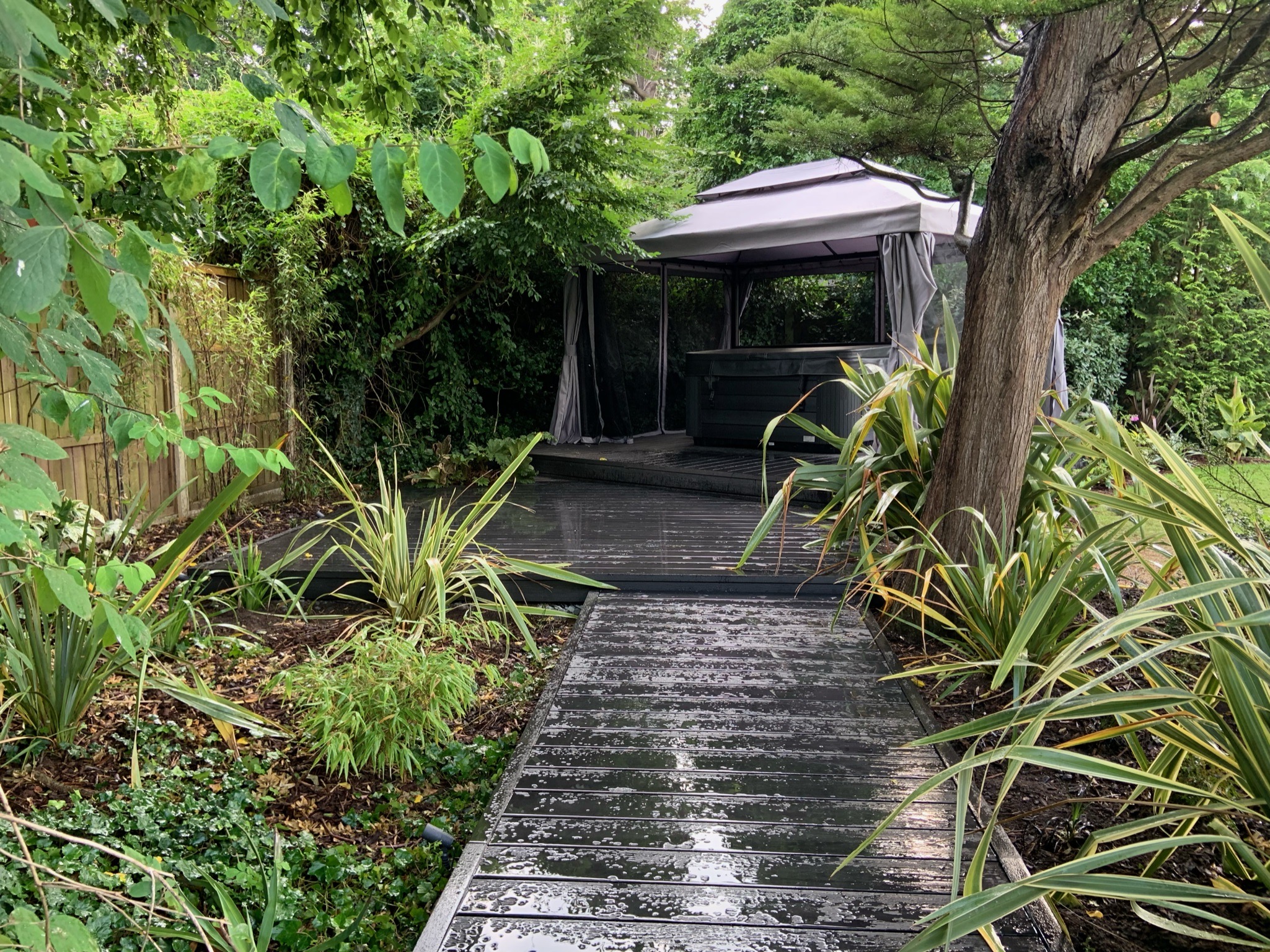 A backgarden featuring a dark reflective wooden walkway leading to a sheltered grey garden gazebo designed by Willow Alexander