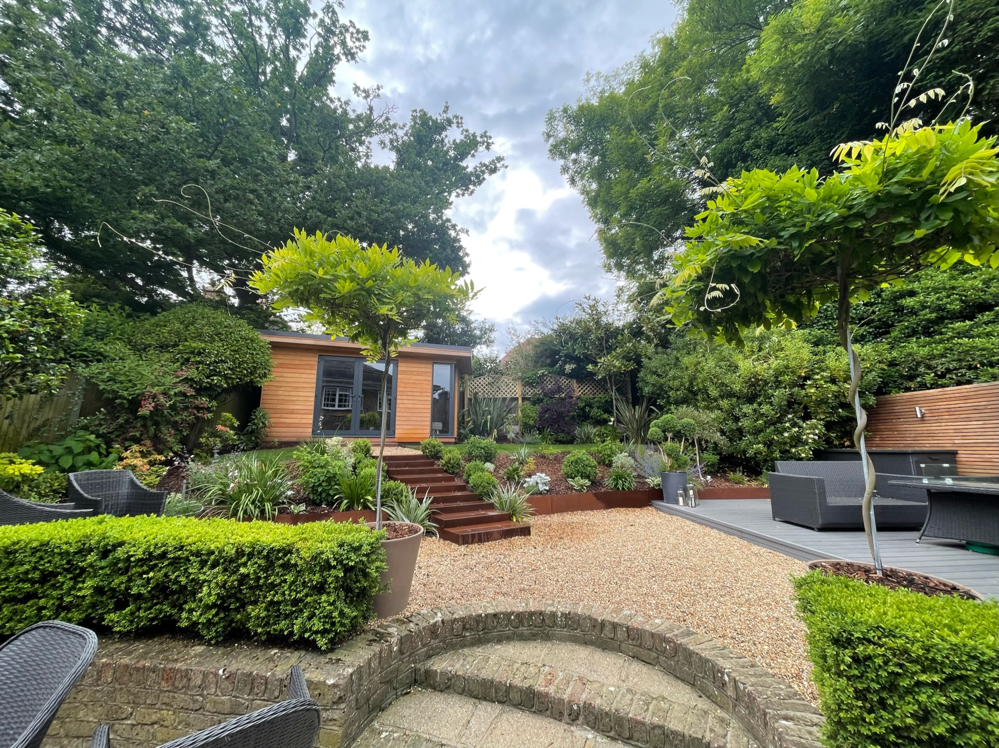 A backgarden with steps leading up to a gravel floor patio connected to a tile floor patio with a rattan furniture set and a garden shed designed by Willow Alexander