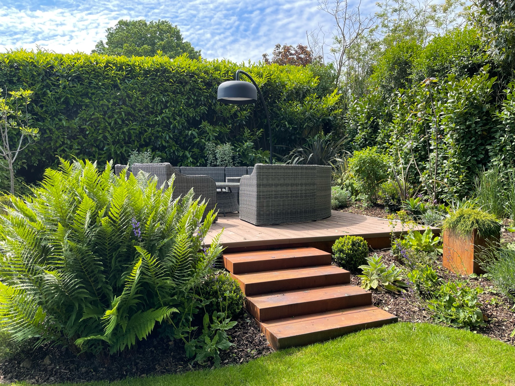 A well-maintained garden with a raised wooden platform with a multi-seating rattan garden furniture set surrounded by high hedges designed by Willow Alexander