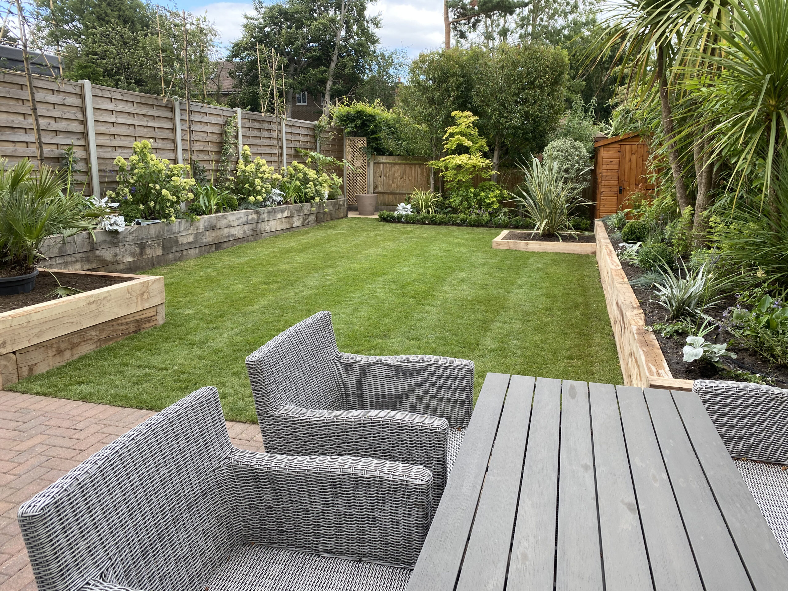 A brick tiled patio with rattan armchairs around a garden table leading out into a well-maintained garden law surrounded by wooden box planters designed by Willow Alexander