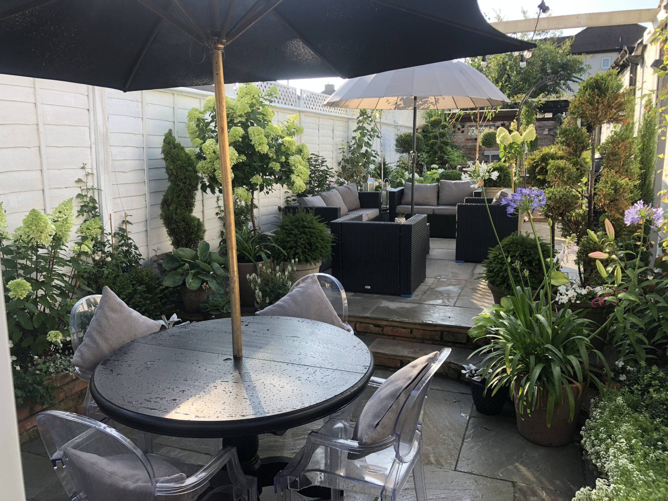 A multi-tiered tiled garden patio filled with potted plants surrounding a glass top garden table with an umbrella and a seven-seater rattan furniture set designed by Willow Alexander