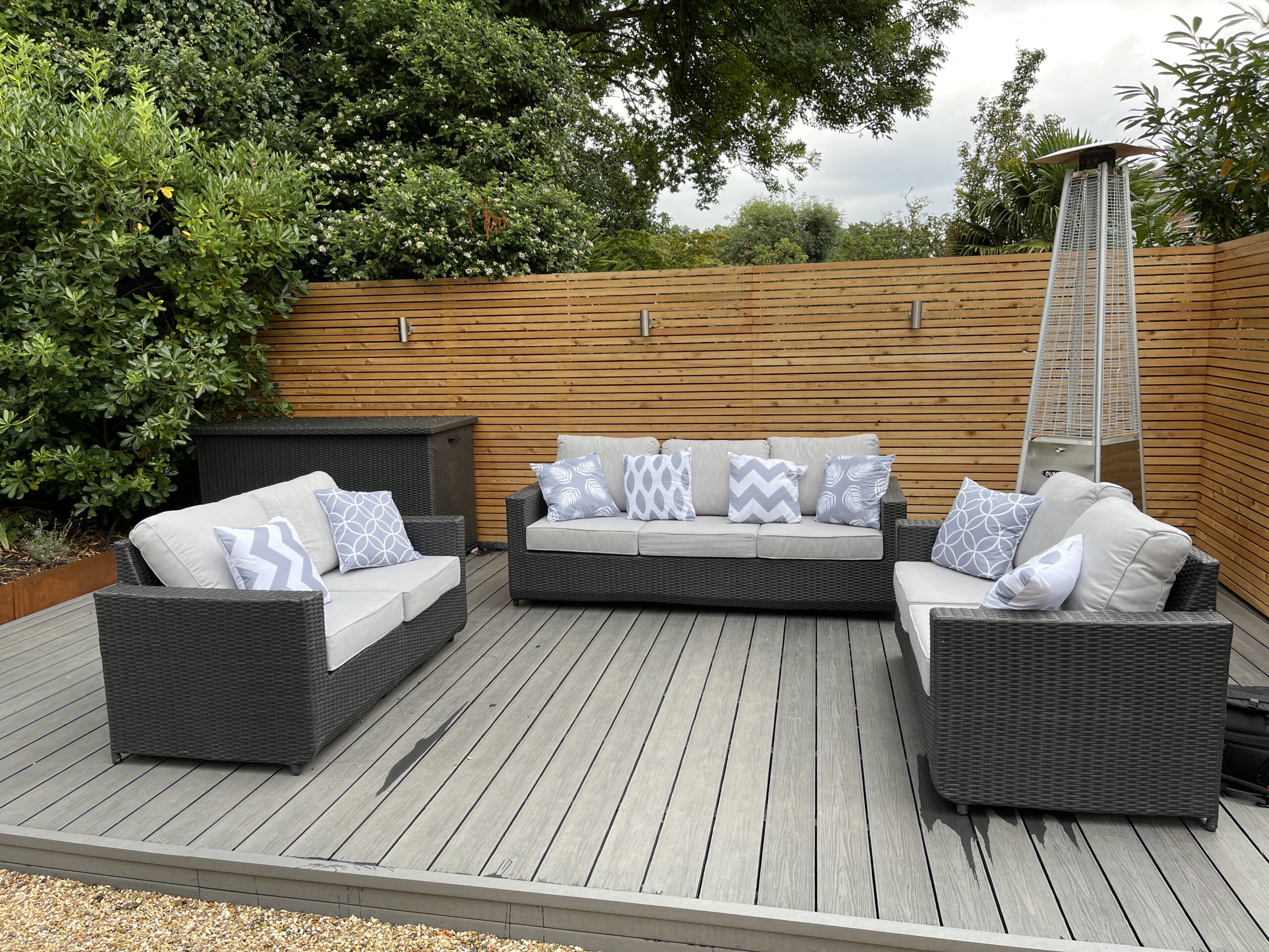 A grey wooden tiled patio with a 9-seater rattan garden furniture set at an outdoor heater surrounded by a modern wooden fence designed by Willow Alexander